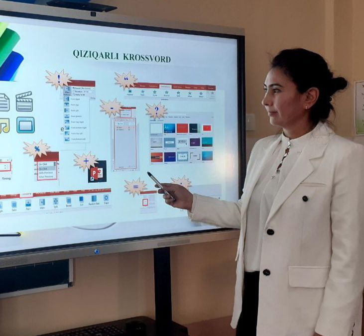 Central Asian woman with dark hair up in a bun and a white coat stands in front of a computer screen pointing out parts of a chart with a pen.