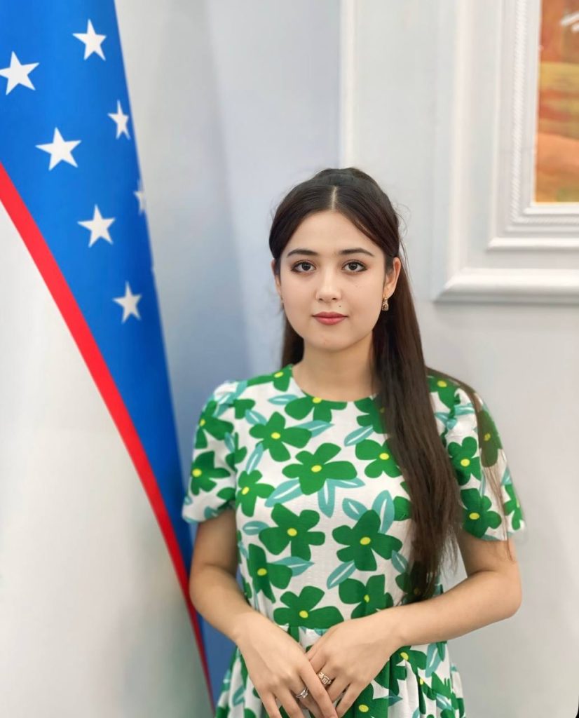 Young Central Asian woman with long dark hair and a dress with a green flower pattern standing next to an Uzbek flag.