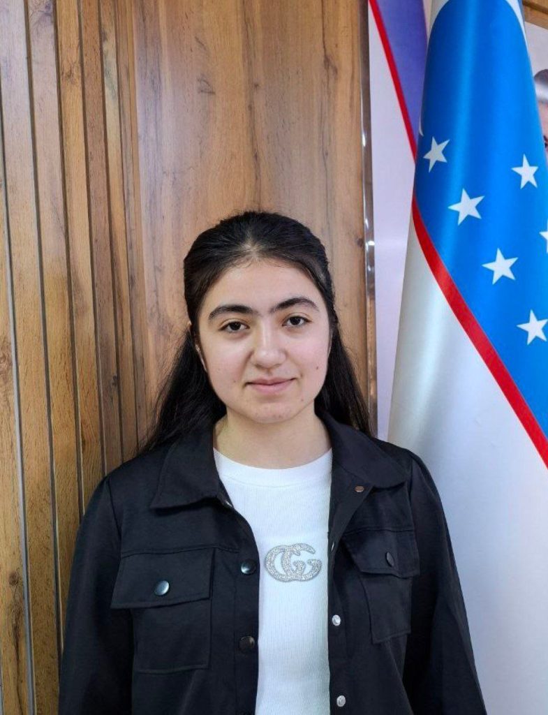 Young Central Asian woman with long straight dark hair, dark coat, and a white tee shirt standing next to a wooden wall and the Uzbek flag.
