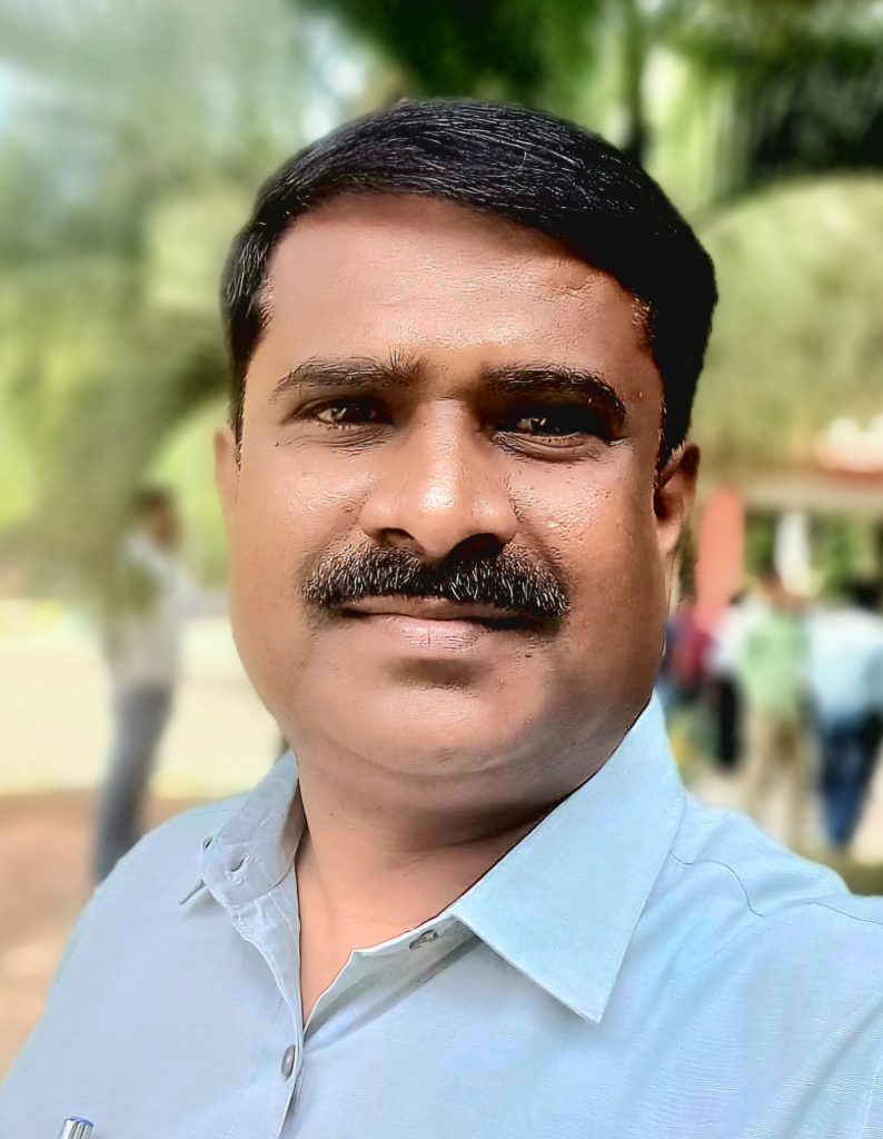 Middle aged South Asian man in a light blue collared shirt. He's got short hair and a trimmed mustache and is outside on a sunny day with trees and other people behind him.