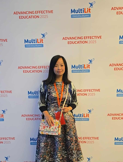 East Asian young woman with long dark hair, colorful floral dress, and purse and lanyard standing in front of a wall with "Advancing Effective Education" printed on it.