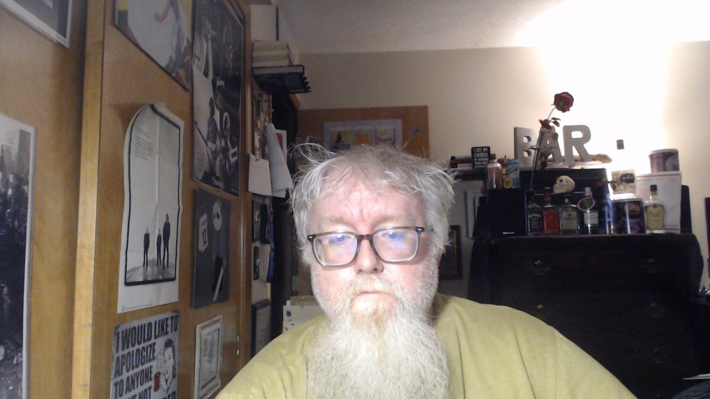 Middle aged white guy in a light colored tee shirt with a long white beard and mustache and messy gray hair and reading glasses in a bedroom with posters on the wall.