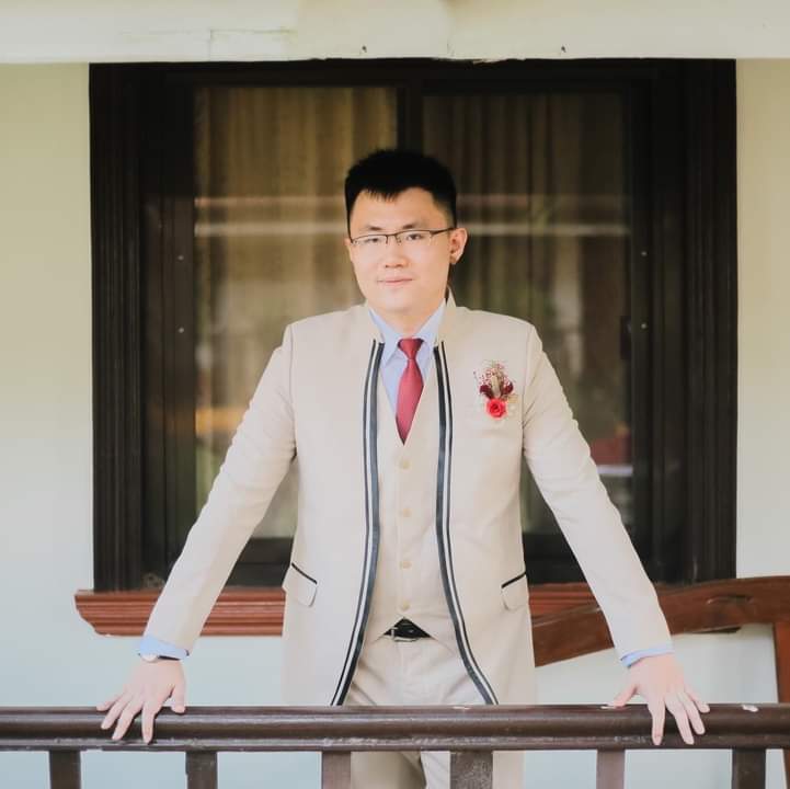 Young East Asian man with short dark hair, clean shaven, with reading glasses, in a tan suit and red tie and corsage.