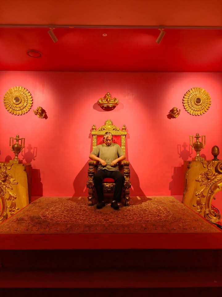 East Asian older man in a short sleeved tan shirt and dark pants seated on an old style carved wooden chair next to other Asian looking museum artifacts on a red wall.