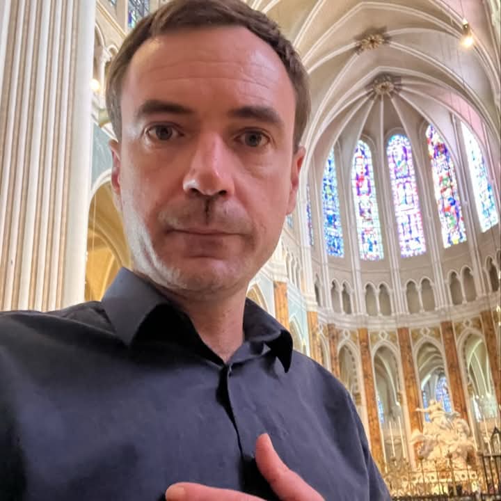 Middle aged European man with short dark hair, clean shaven, in a blue collared dress shirt inside a white cathedral with stained glass windows on a sunny day.