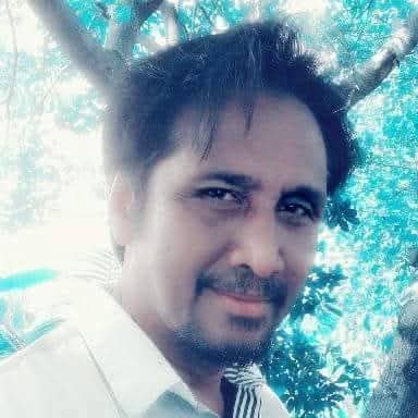 Asian Pacific Islander man with trimmed beard and mustache, short brown hair and brown eyes, and a white collared shirt standing in front of a leafy tree.