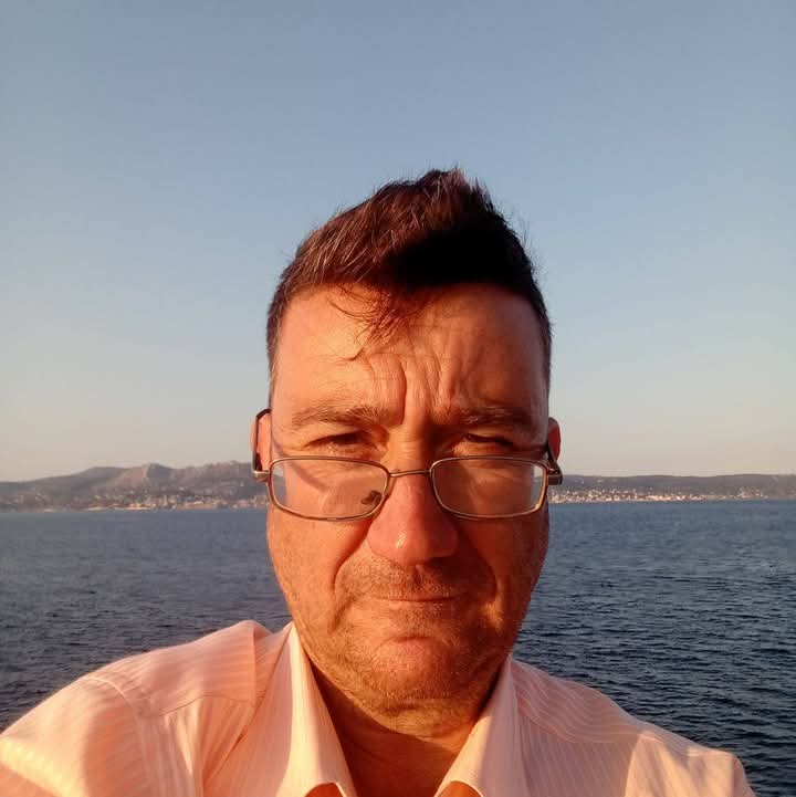 Middle aged European man with reading glasses and a light colored collared shirt in front of water.