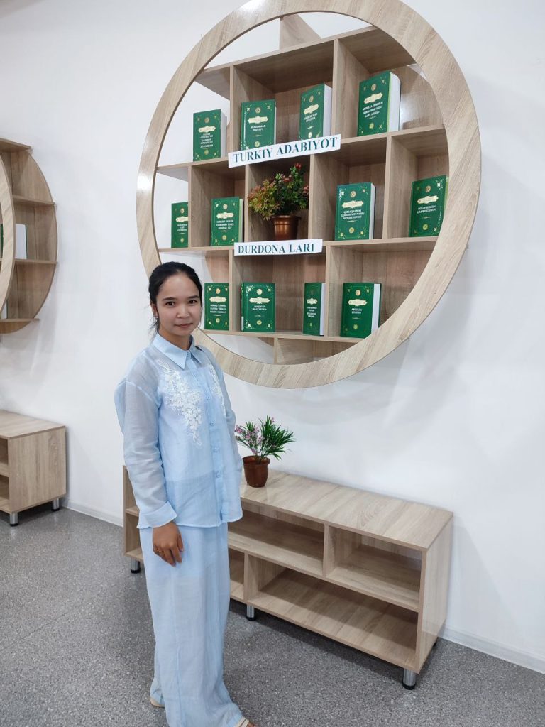 Young Central Asian woman with dark hair and a white collared shirt in front of a bunch of books with green covers on a wooden shelf. She's in a white dress shirt and white pants.