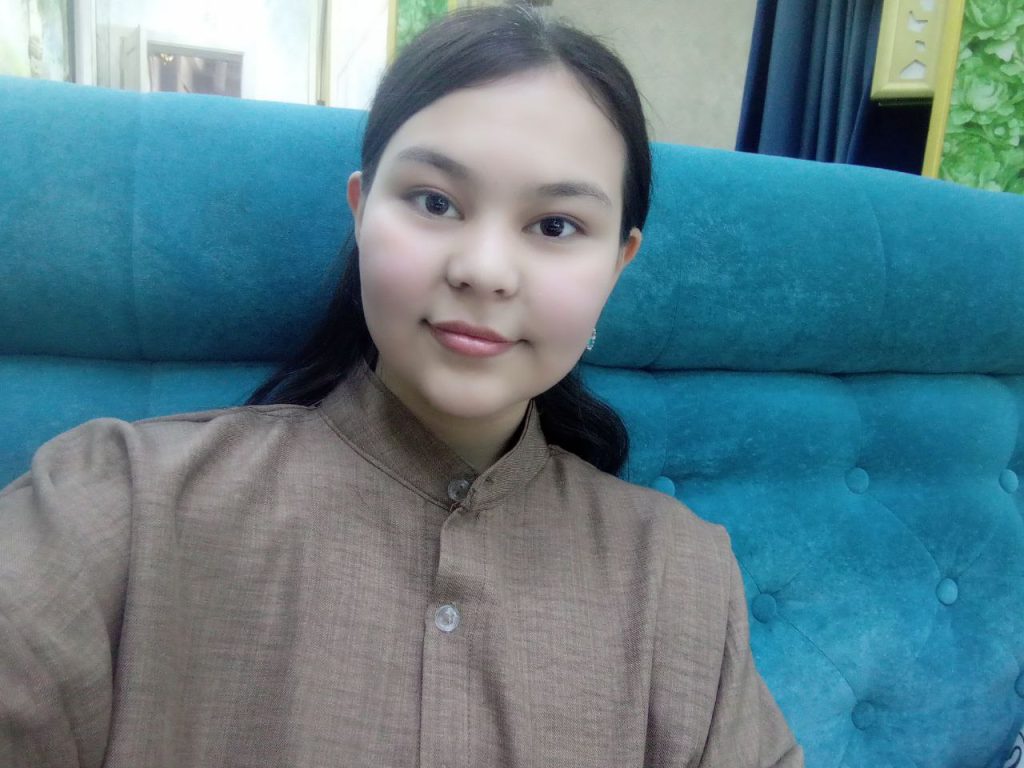 Young Central Asian woman with straight dark hair and brown eyes and a light tan blouse seated on a blue couch