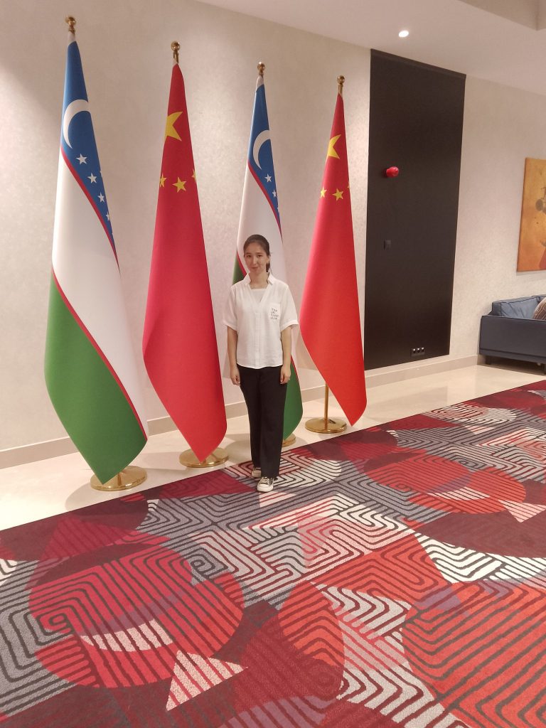 Young Central Asian woman in a white collared shirt and black pants standing in a public building on a carpet in front of a bunch of flags.