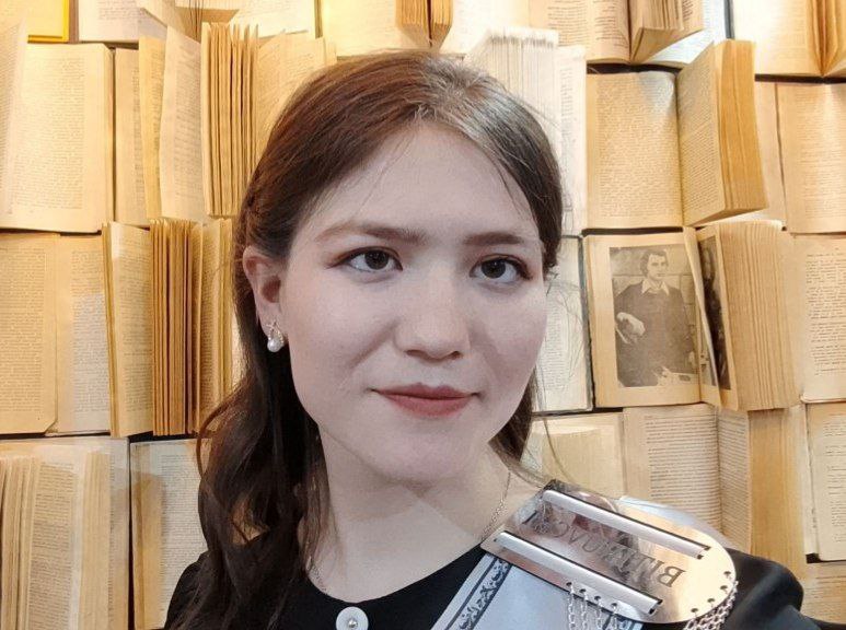 Central Asian teen girl with long straight dark hair, brown eyes, small white earrings, and open paperback books behind her.