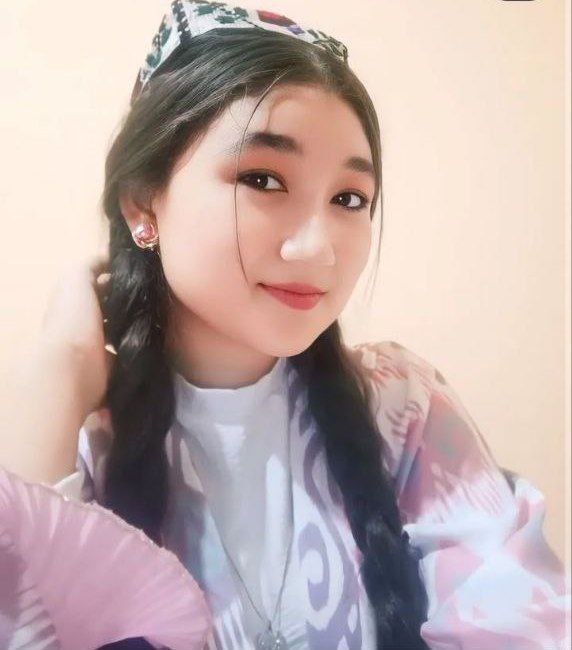Central Asian girl with dark braids, brown eyes, and a smile, in a white top and a pink patterned sweater.