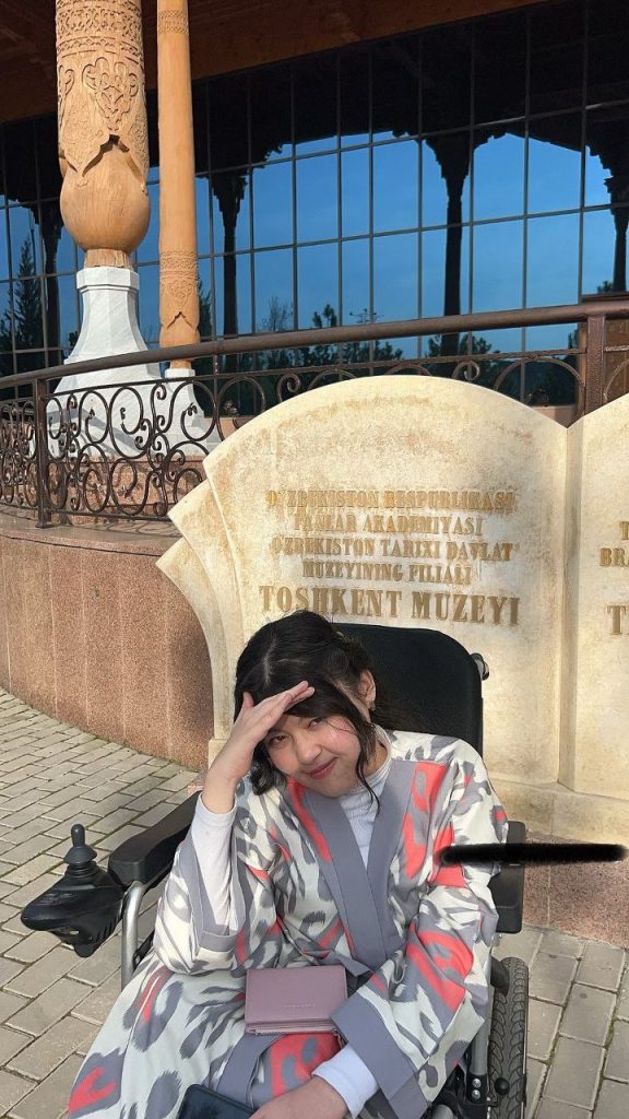 Young Central Asian woman with a light gray, white, and red coat seated in a chair next to a stone monument to an important person.