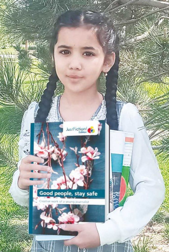 Young Central Asian girl with dark pigtails and a white blouse and blue overalls holding a set of books in front of a pine tree on a grassy field on a sunny day.