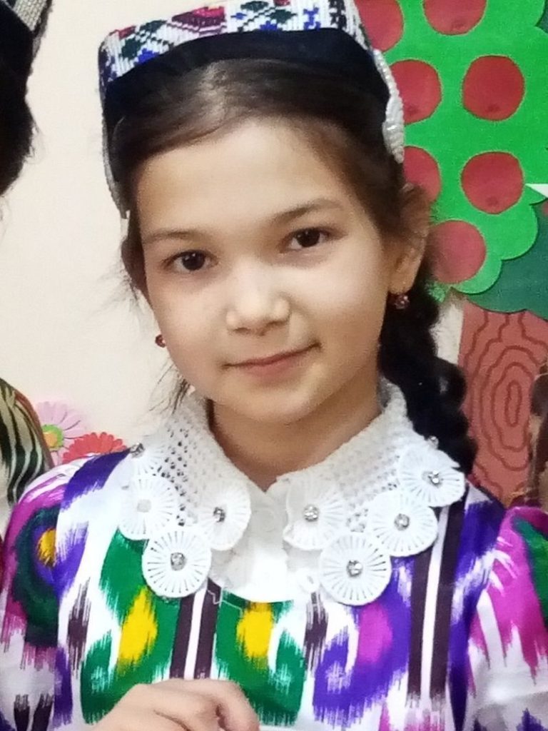 Young Central Asian girl with an embroidered headband and colored blouse in front of a construction paper mural.
