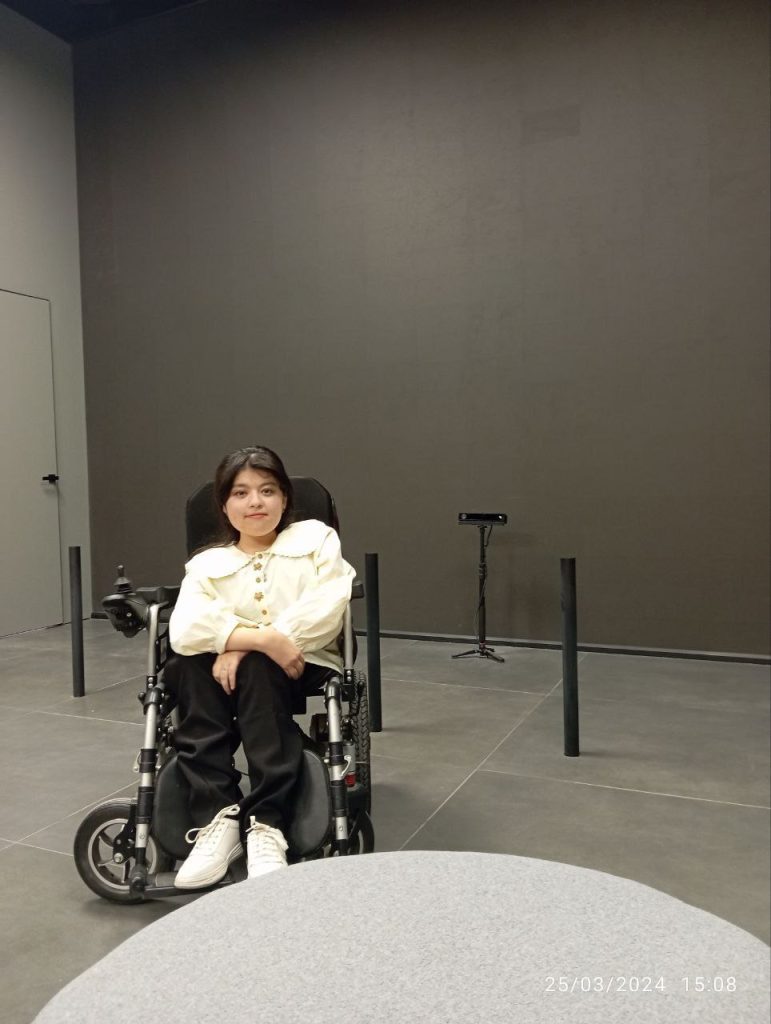 Dark haired Central Asian woman with a white collared blouse and black pants and white tennis shoes in a wheelchair.