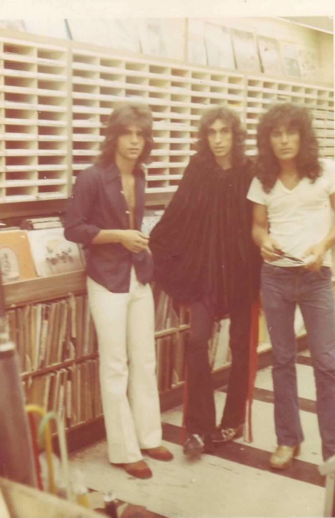 Three young white adults with poofy 70's hair and big collared shirts and long jeans standing in front of records on display in a store