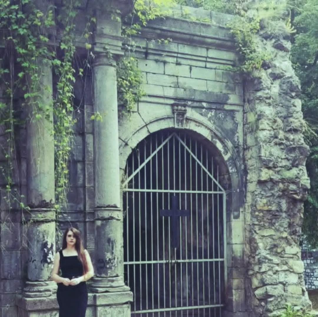 Young European woman with long dark hair and a black dress stands near a green mossy stone castle entrance. Stones are draped with ivy and she's a small figure in the lower left.