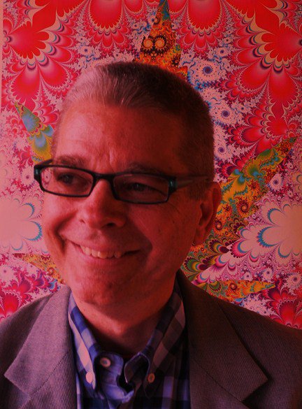 Middle aged white man in reading glasses and a gray suit and blue and white checkered shirt with short gray hair in front of a red and white and yellow and blue and green psychedelic fractal design background.