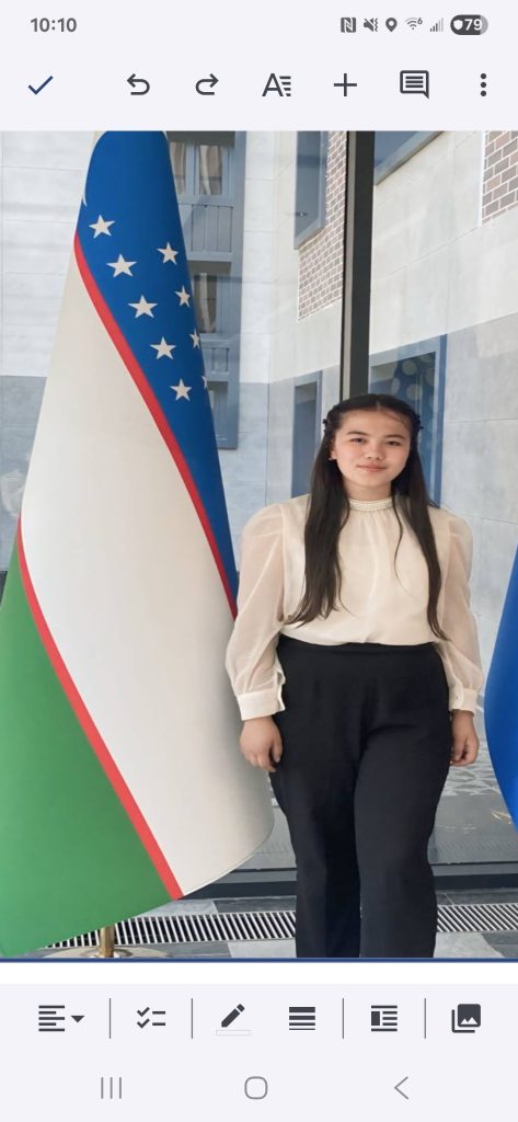 Young Central Asian woman with long dark hair, a white blouse, dark pants, standing in a room with big windows and the Uzbek flag.