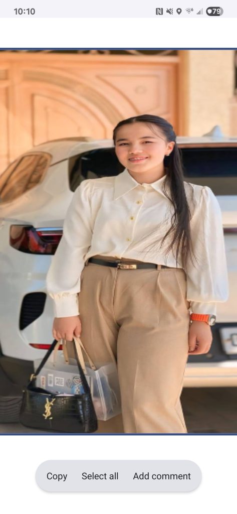 Young Central Asian woman with long dark hair and a collared off white blouse and a belt and tan pants, holding a purse in front of a white car. 