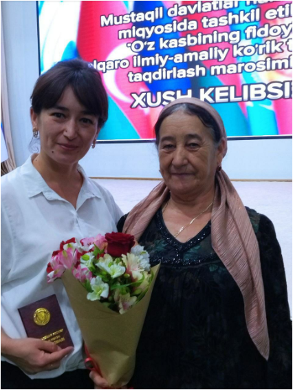 Younger Central Asian woman with dark hair and a white collared shirt standing next to an older Central Asian woman with a pink headscarf and black blouse. The older woman holds a bouquet of flowers.