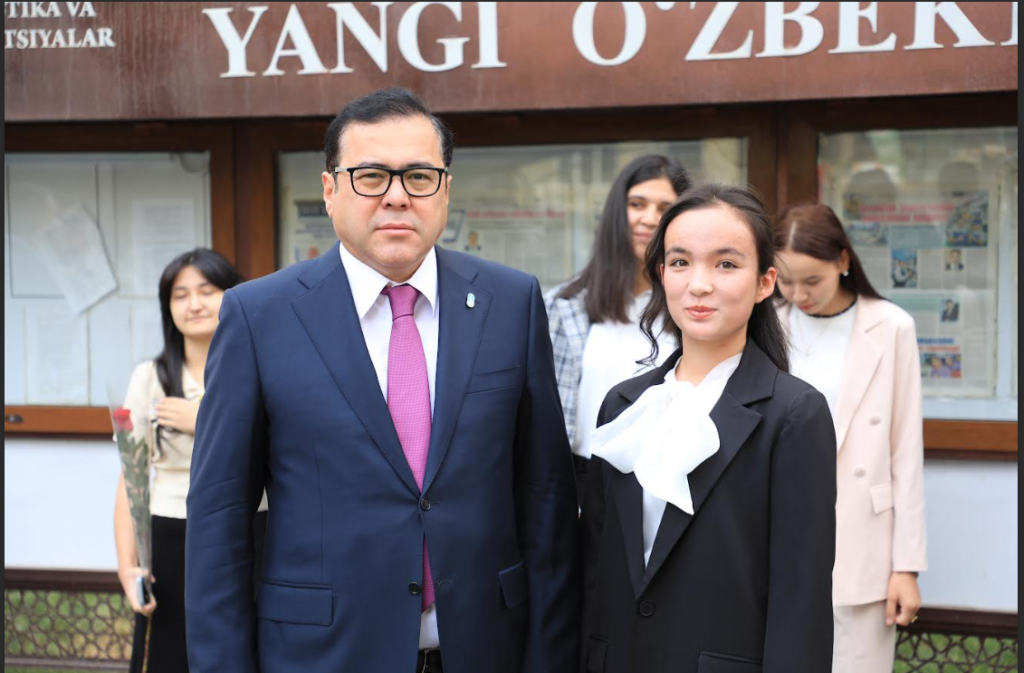 Young Central Asian woman, right, in a white top and black coat, next to a Central Asian man in glasses and a black suit and red tie. Other young women in the background, bulletin board in background behind all of them.