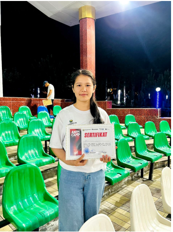Young Central Asian woman holding a certificate. She's in a white tee shirt and blue jeans in a room with empty green chairs.