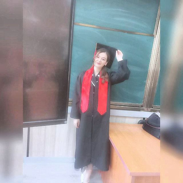 Young Central Asian woman with a red sash and black graduation cap and gown posing in front of a green blackboard.