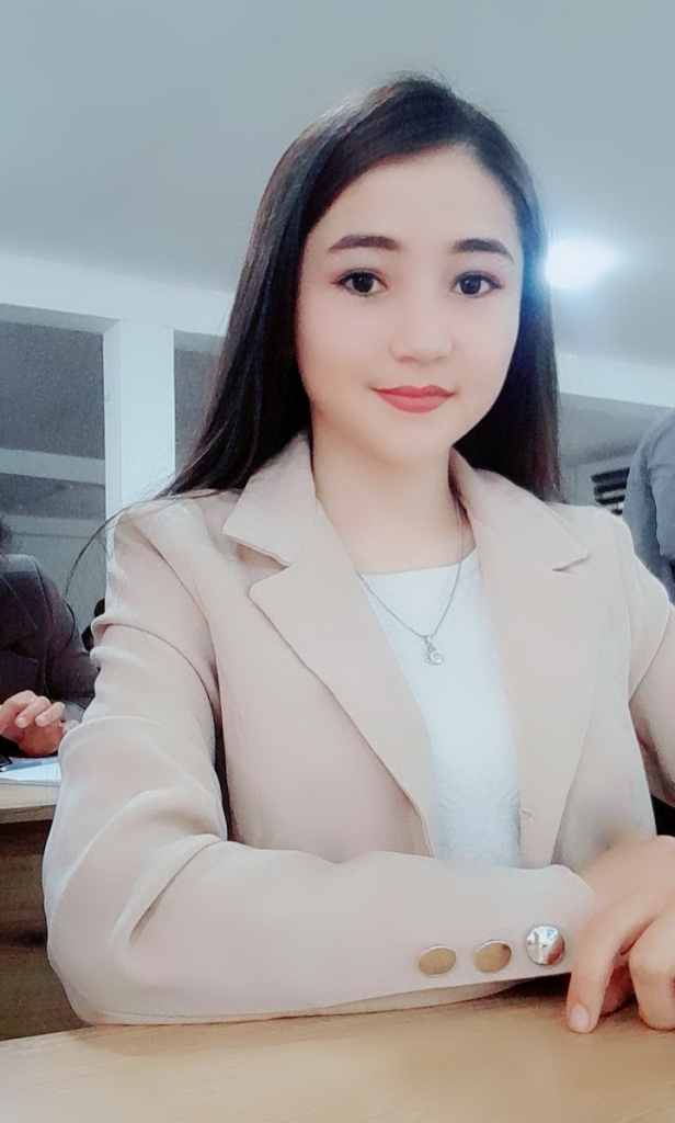 Young Central Asian woman with long straight dark hair, brown eyes, a tan coat, white blouse, and small necklace.