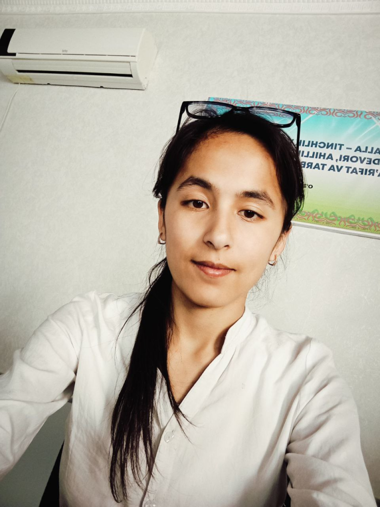 Young Central Asian woman with long dark hair, small earrings, reading glasses on her head, and a white blouse.