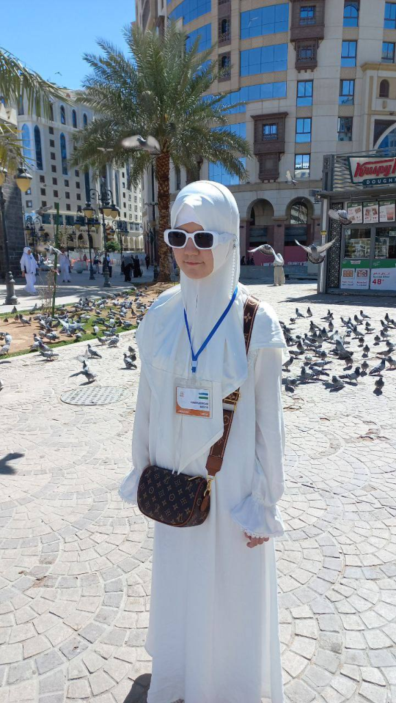 Young Central Asian woman in a white outfit that covers her body and hair. She's got sunglasses and a purse and a lanyard and is standing in a plaza with shops and a lantern and pigeons.