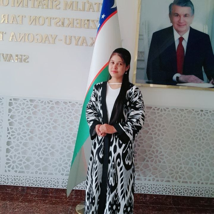 Young Central Asian woman in a long black and white patterned coat and white top, long dark hair. She's in front of a flag and a photo of a man in a black coat and red tie.