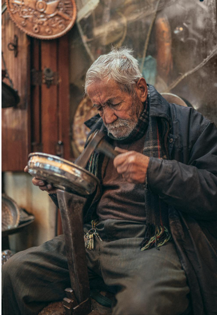 Elderly light skinned gentleman of undetermined race looking deeply into a metal bowl as he shapes the metal with a hammer. He's in a shop with wooden walls and doors and tools.