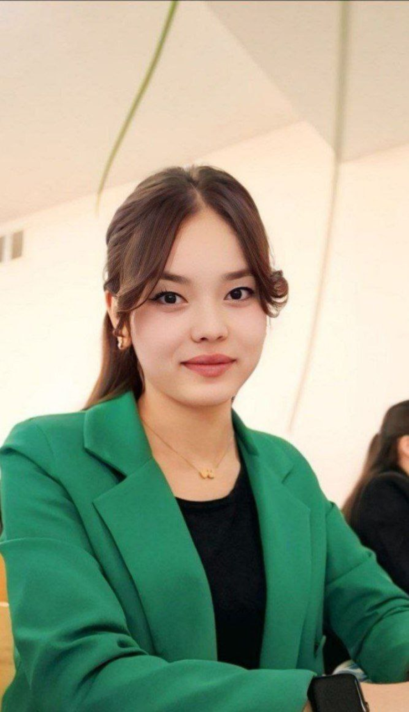 Young Central Asian woman with dark straight hair, brown eyes, and a green coat over a black top. Light colored necklace.