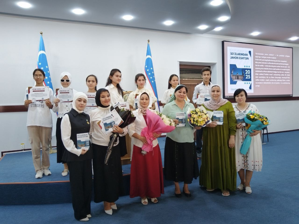 Group of young Central Asian adults in front of Uzbek flags and holding bouquets of flowers and certificates on a small stage in a classroom.