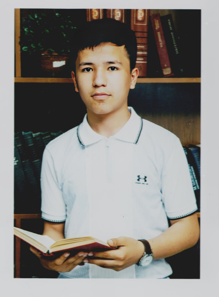 Young Central Asian teen boy in a white collared shirt with short black hair standing in front of a bookshelf. 