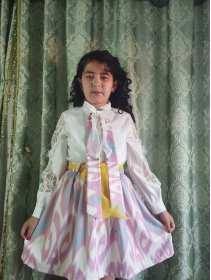 Central Asian teen girl with curly dark hair and a pink and white ruffled dress with a yellow sash in front of a green and white curtain.