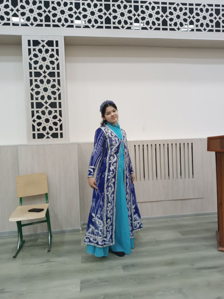 Central Asian teen in a long embroidered blue coat with silver designs and trim and a light blue dress and a headband in a room with Islamic style decorative architecture.