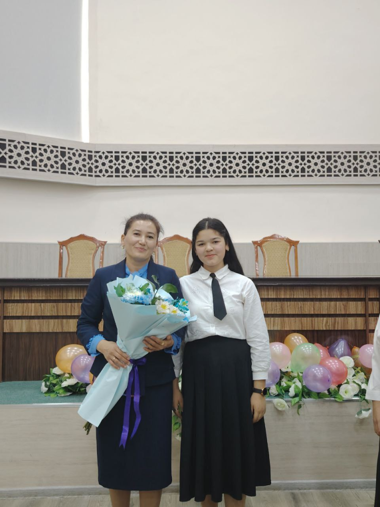 Young Central Asian teen in a white blouse, black tie, and black skirt. She's in a room with chairs and a stage and flowers and balloons. A woman is next to her holding flowers.