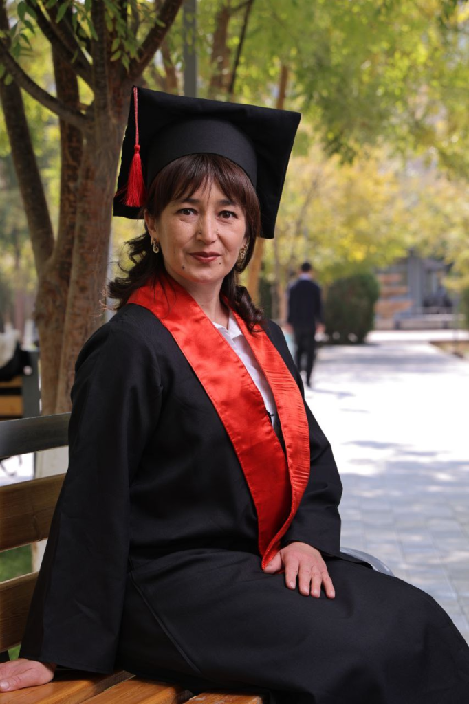 Central Asian woman with straight dark hair dressed in a black graduation robe and hat with a red sash and tassel. She stands outside in front of a leafy tree on a concrete path. 