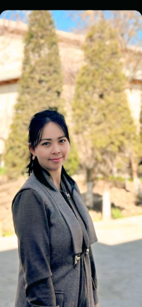 Young Central Asian woman with straight dark hair and a gray coat in front of some conifer trees and a building on a sunny day.