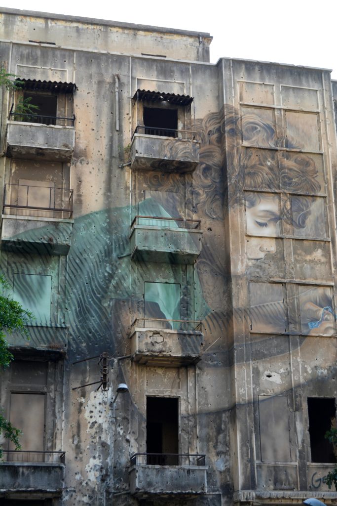 Painting of a curly haired small child with a green top engrossed in play on the wall of a gray apartment building (Lebanon)
