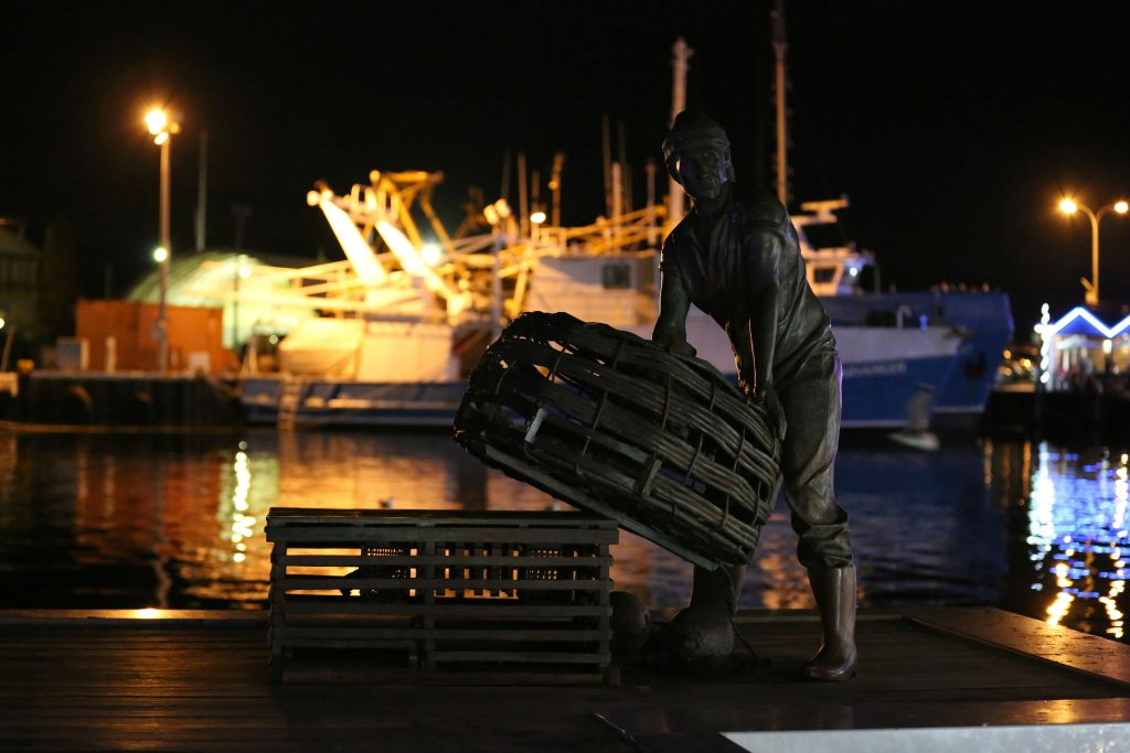 Dark metal statue of a man in rolled-up pants holding a large wicker basket over a wooden crate at night near water, a large ship, and a pier. (South Asia)