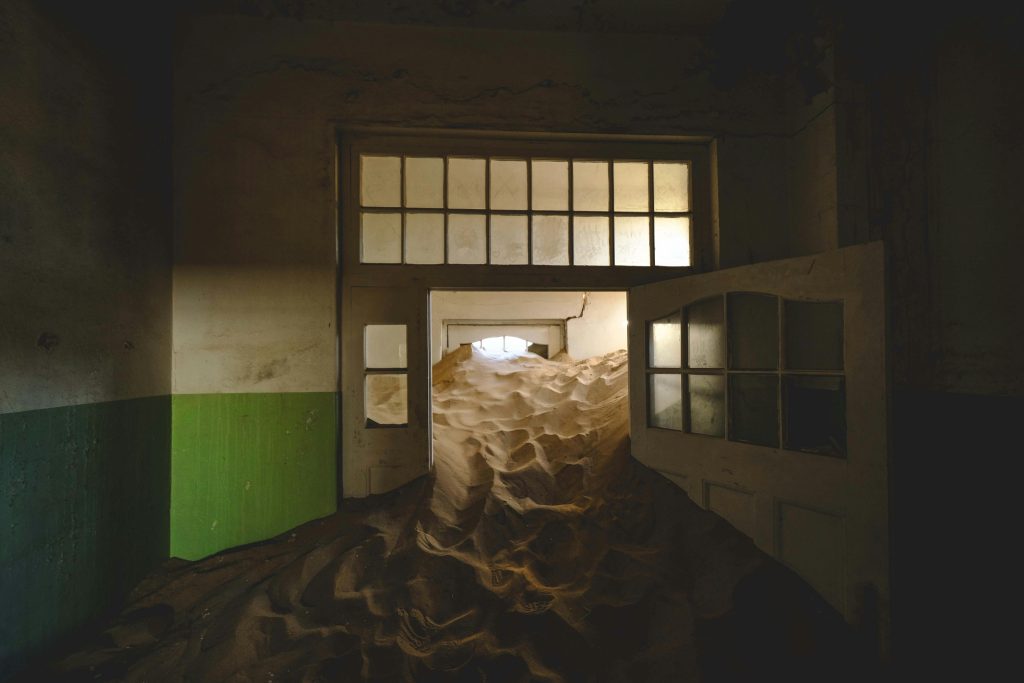 Artsy photo of sand in a doorway in an abandoned room with sunlight streaming in through windows. White walls with green paint (Namibia)