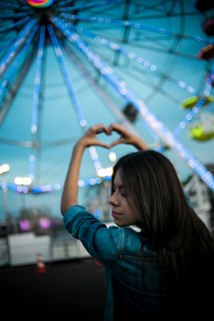 Woman making a heart symbol with her hands in front of a lit carnival ride at dusk. She's got long dark hair and is in a jean jacket.