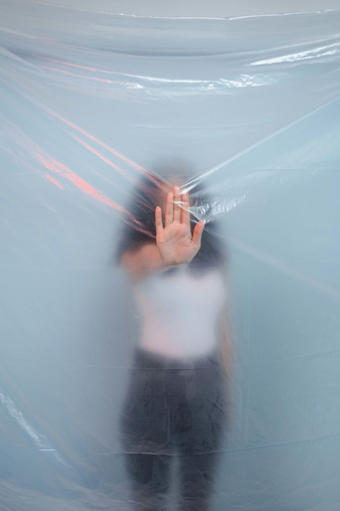 Light skinned woman puts her hand in front of her face reaching out against plastic that partially obscures her. She's got dark hair and a white blouse.