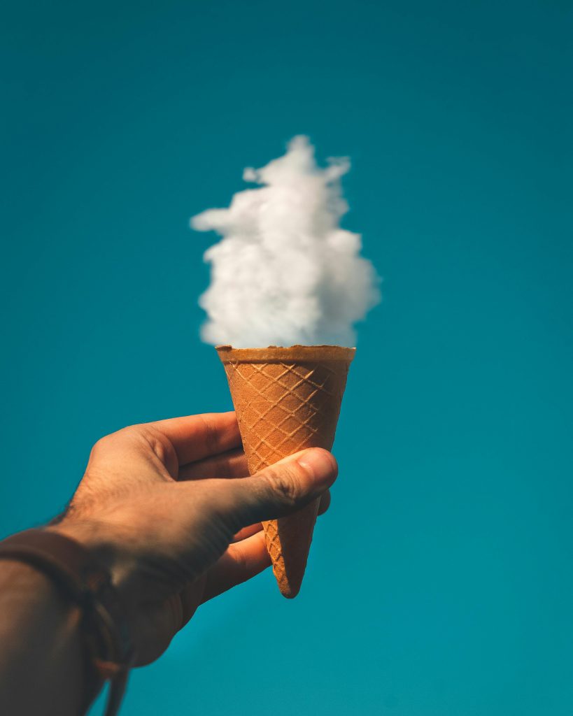 Fanciful image of a white fluffy cloud in an ice cream cone held in a person's hand in a blue sky.