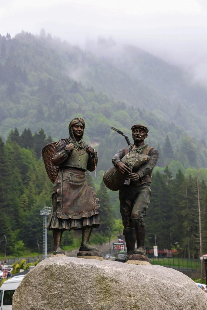 Light brown and green metal statues of traditionally dressed Turkish man and woman. Caps, headscarves, vest and dress and petticoat and boots. They're in a misty conifer forest.