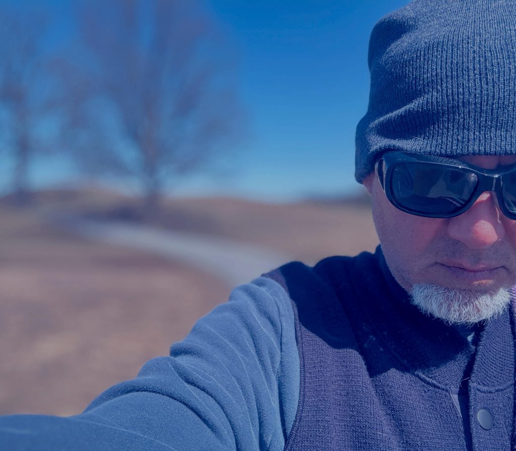 Middle aged white man with dark sunglasses, a knit hat, a small trimmed beard, and a dark sweater on a hiking train with a path and some barren trees on a sunny day.